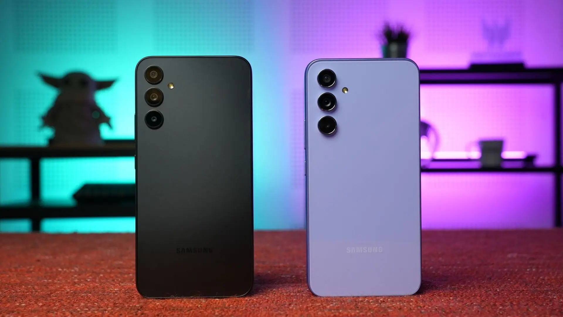 Two Samsung smartphones shown from the back on a red surface, black and lavender finishes with multiple rear cameras and a neon backdrop.