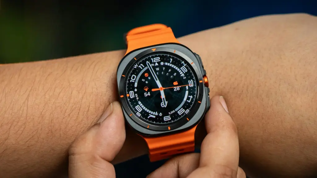 Close-up of an orange-strapped smartwatch on a wrist, displaying a busy analog-digital watch face with orange accents.