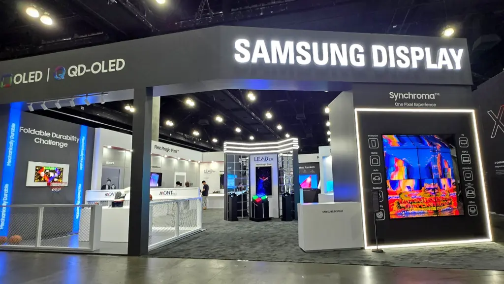 Trade show booth labeled Samsung Display with OLED and QD-OLED signs; large TV wall and product displays in a dark exhibition hall.