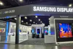 Trade show booth labeled Samsung Display with OLED and QD-OLED signs; large TV wall and product displays in a dark exhibition hall.