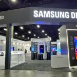Trade show booth labeled Samsung Display with OLED and QD-OLED signs; large TV wall and product displays in a dark exhibition hall.
