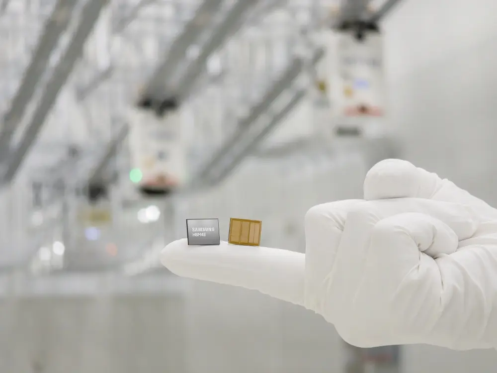 Gloved hand holds two small computer chips, one grey labeled 'SAMSUNG HBM4E', in a pristine cleanroom backdrop with blurred industrial equipment.