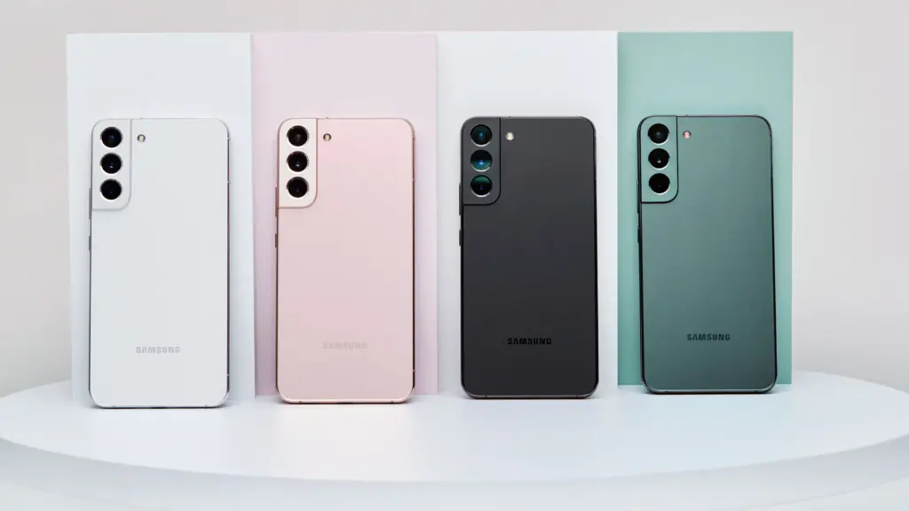Four Samsung smartphones shown from the back in white, pink, black, and green, standing on pastel panels on a white surface.