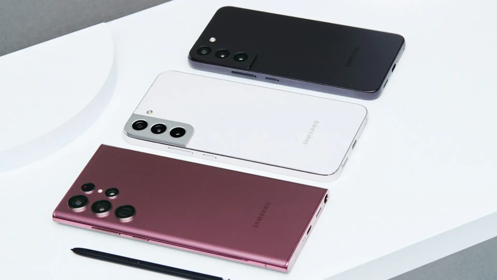 Four Samsung Galaxy phones in black, white, burgundy, and pink arranged diagonally on a white surface, with an S Pen in the foreground.