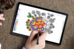 Tablet on carpet, hand using a stylus to color a floral design in a digital coloring app; color palette along the edge is visible.