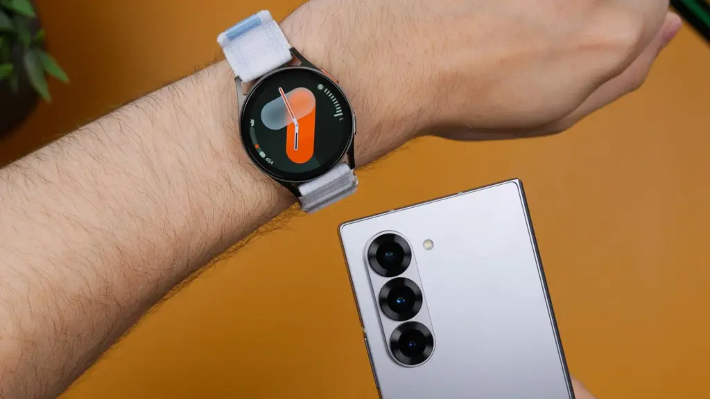 Round smartwatch with a light gray strap on a hairy forearm, next to a silver smartphone with multiple rear cameras on an orange background.
