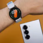 Round smartwatch with a light gray strap on a hairy forearm, next to a silver smartphone with multiple rear cameras on an orange background.