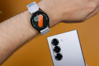 Round smartwatch with a light gray strap on a hairy forearm, next to a silver smartphone with multiple rear cameras on an orange background.
