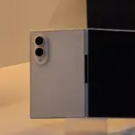Three smartphones on a white display: a white foldable with dual circular camera module on its back, flanked by black devices with blank screens.