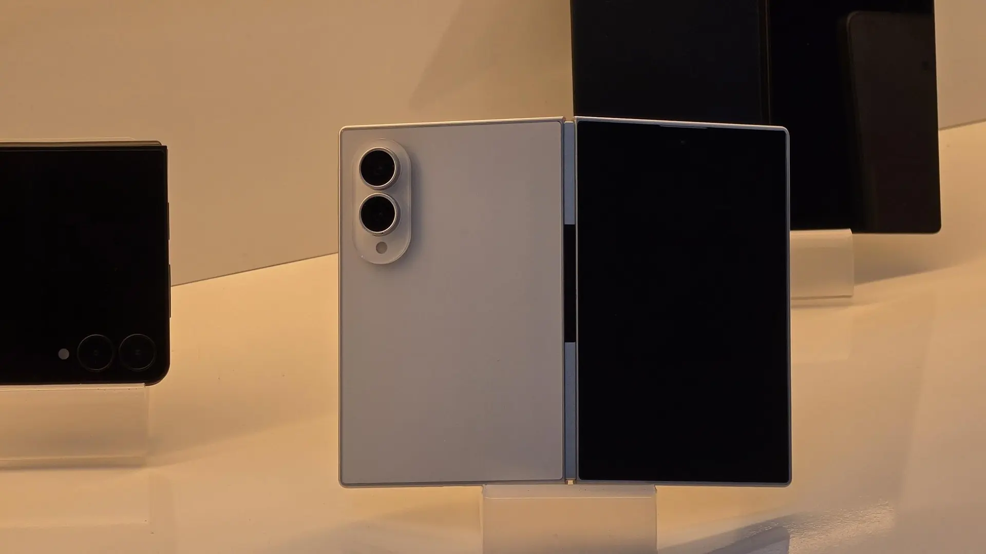 Three smartphones on a white display: a white foldable with dual circular camera module on its back, flanked by black devices with blank screens.