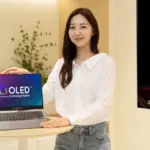 Smiling woman in a white shirt stands beside a round table with a Samsung OLED laptop; a large DisplayHDR ad is on the right edge.