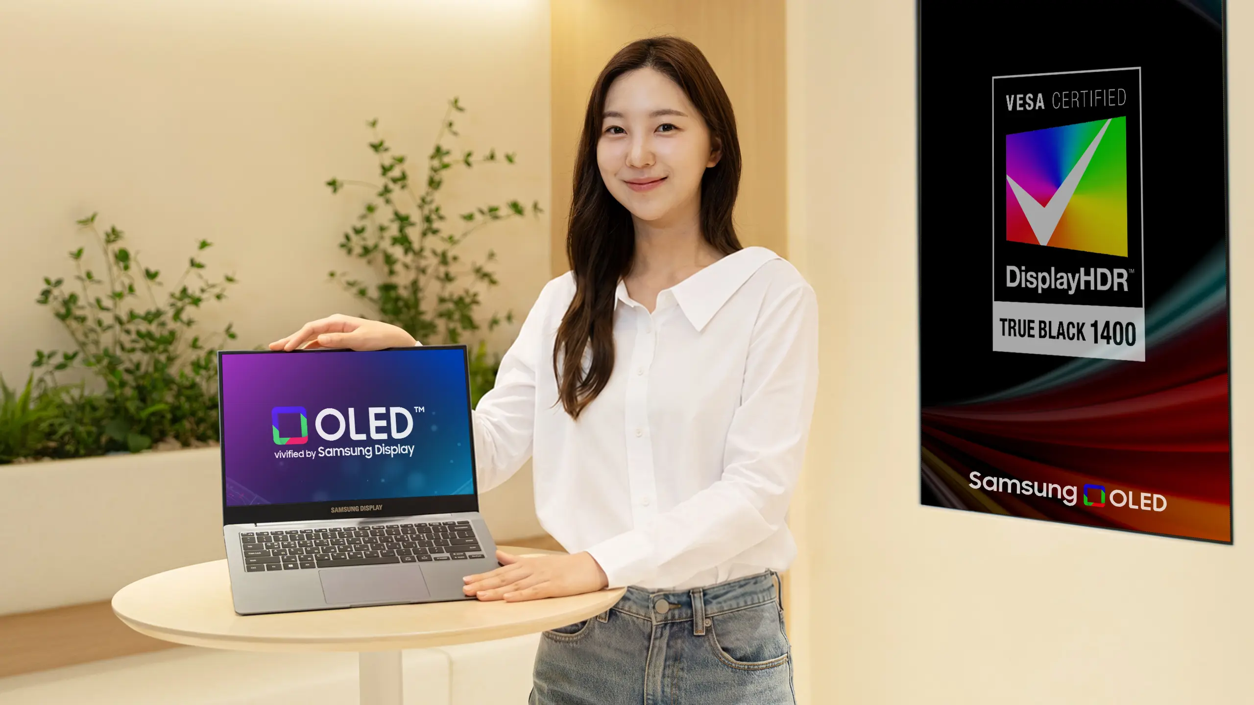 Smiling woman in a white shirt stands beside a round table with a Samsung OLED laptop; a large DisplayHDR ad is on the right edge.
