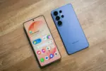 Two modern smartphones lie on a wooden table, a peach-colored front phone showing app icons and widgets next to a blue phone with multiple rear cameras.
