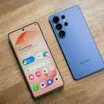 Two modern smartphones lie on a wooden table, a peach-colored front phone showing app icons and widgets next to a blue phone with multiple rear cameras.