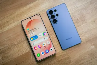 Two modern smartphones lie on a wooden table, a peach-colored front phone showing app icons and widgets next to a blue phone with multiple rear cameras.