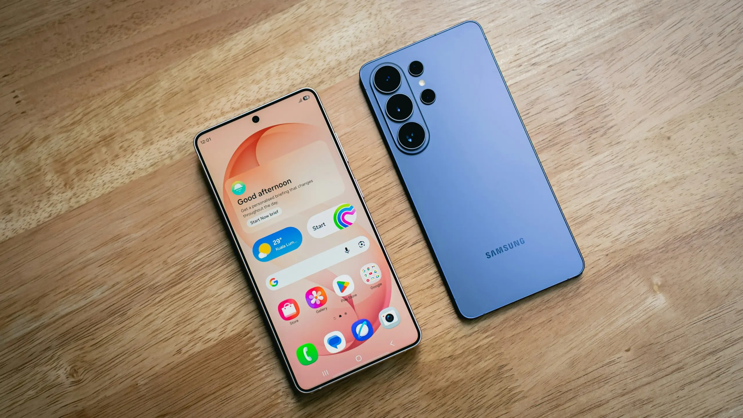 Two modern smartphones lie on a wooden table, a peach-colored front phone showing app icons and widgets next to a blue phone with multiple rear cameras.