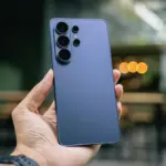 Hand holding a blue Samsung smartphone with a quad-camera setup on the back, outdoor blurred background