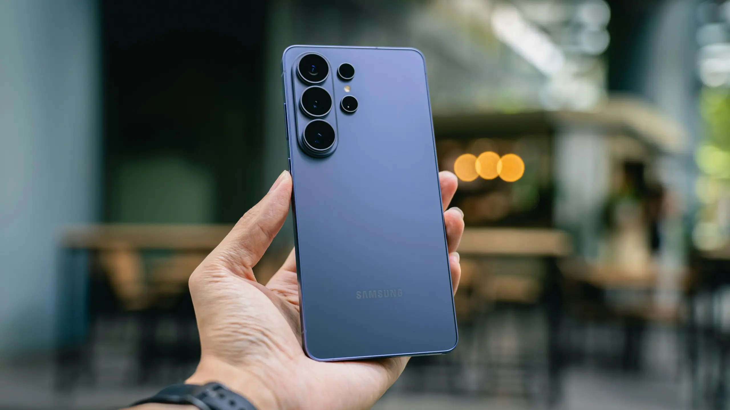 Hand holding a blue Samsung smartphone with a quad-camera setup on the back, outdoor blurred background