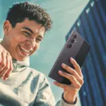 Young man in a gray hoodie smiling while taking a selfie with a large multi-camera smartphone outdoors near a glass building