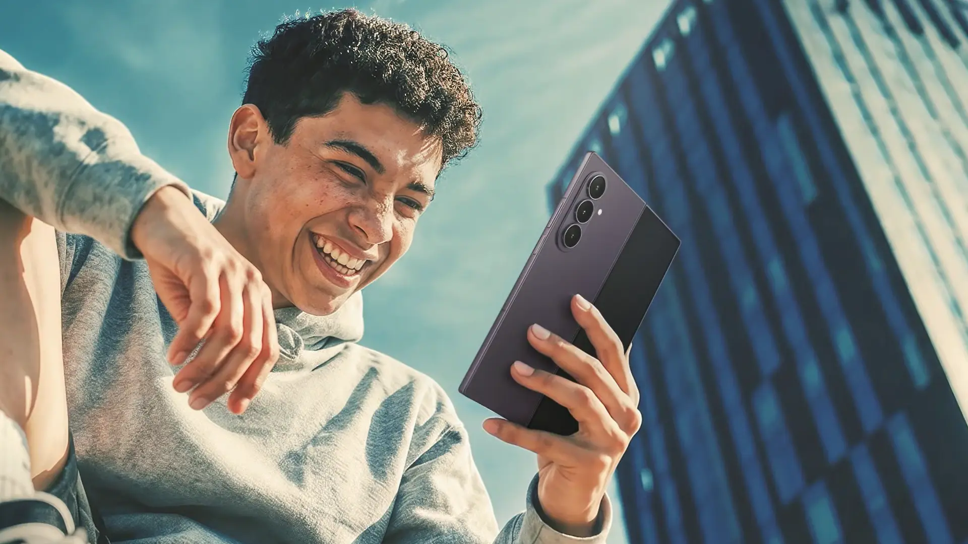 Young man in a gray hoodie smiling while taking a selfie with a large multi-camera smartphone outdoors near a glass building