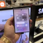 Hand holding a smartphone showing quick settings (Wi‑Fi, Bluetooth) on the screen in a Samsung store display background.