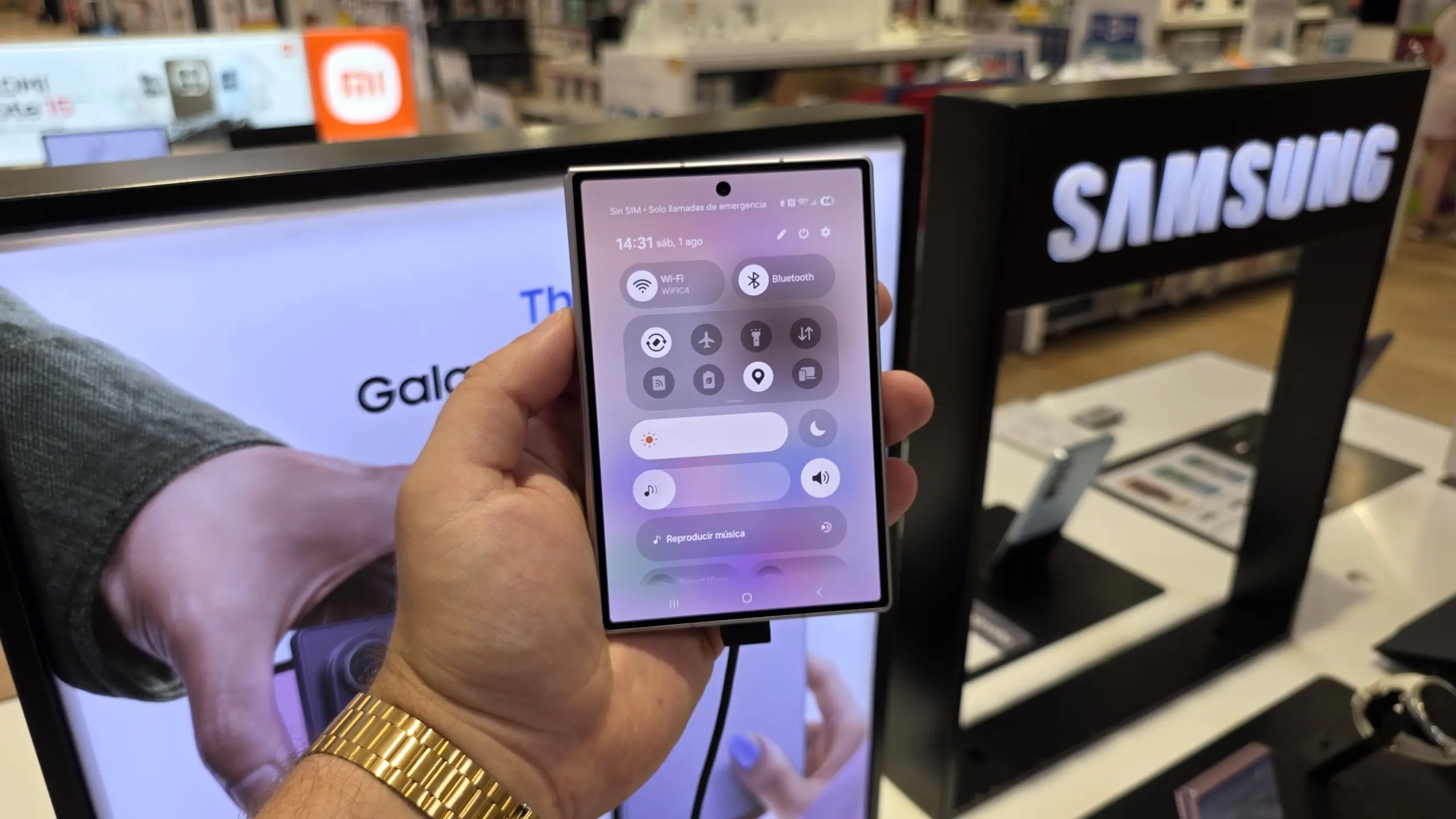 Hand holding a smartphone showing quick settings (Wi‑Fi, Bluetooth) on the screen in a Samsung store display background.